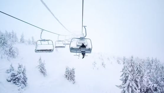 Image of a ski lift in the snow. There is one person riding and looking out over the snow covered treetops.