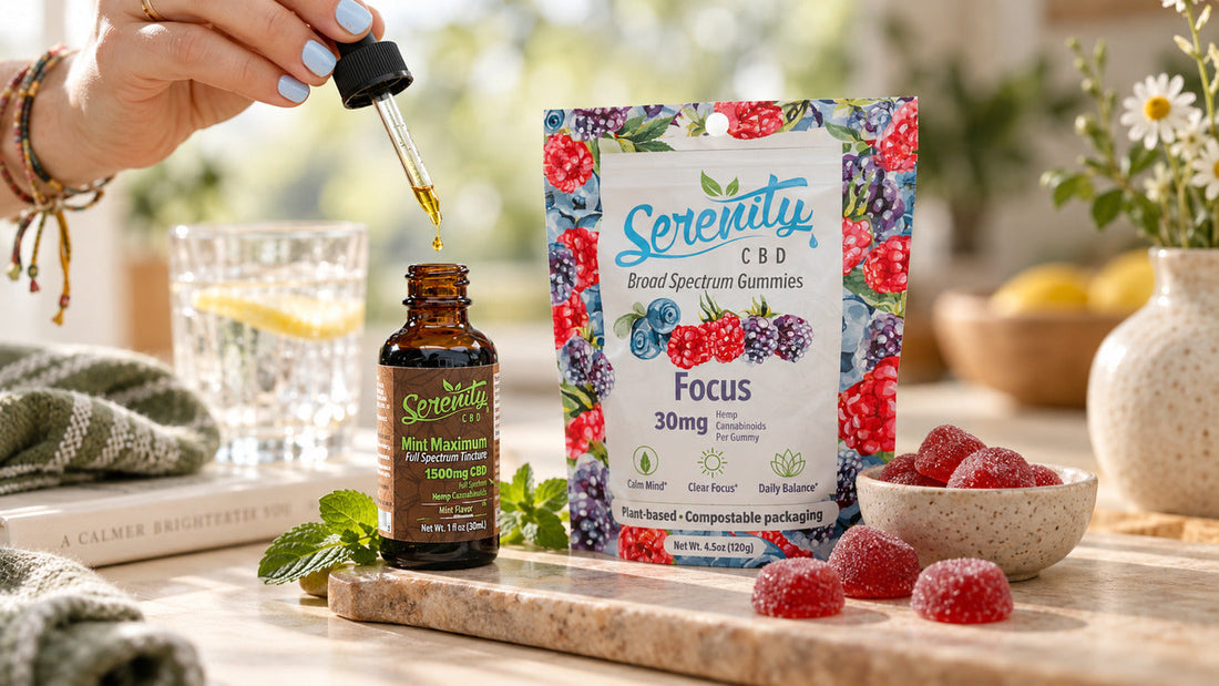 Serenity CBD products on a wooden board: hand with light blue nails dripping oil from a tincture bottle next to a package of Focus Broad Spectrum Gummies and a bowl of red gummies, with a glass of lemon water and fresh mint in a bright kitchen setting