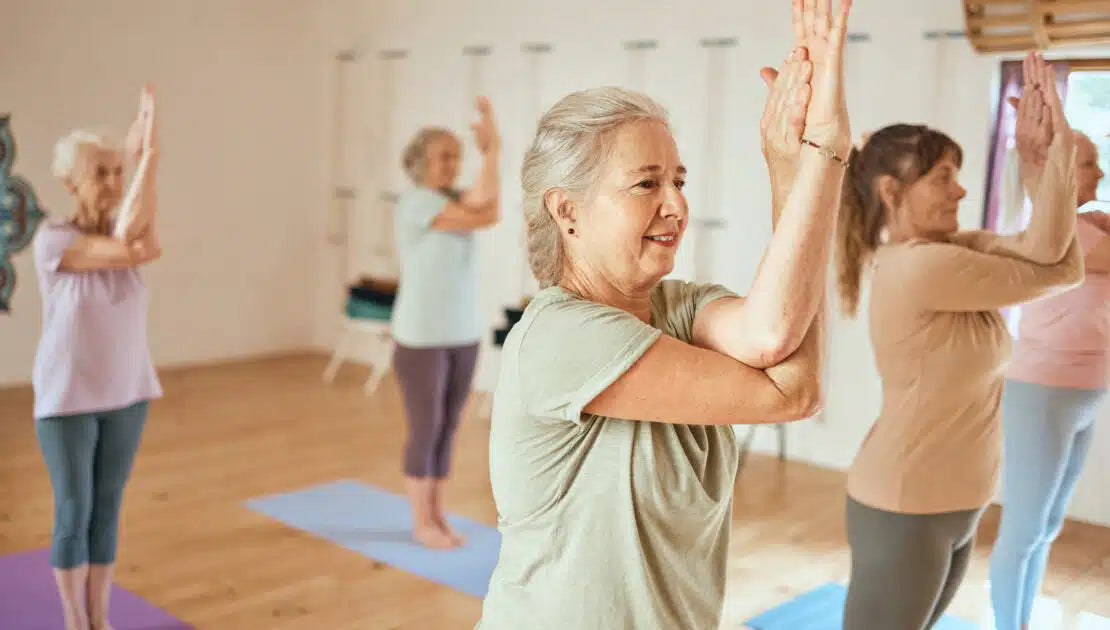 A class of older women practicing yoga. They all have their hands clasped together as they stand on blue mats.