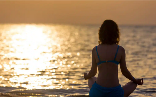 Image of the back of a woman overlooking a sunrise sitting on a yoga mat ready to start the day with meditation.