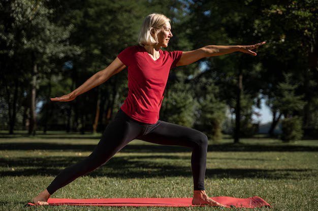 A mature woman practices a pose to build core strength with yoga in a park on a red mat, wearing a red shirt.