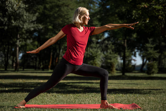A mature woman practices a pose to build core strength with yoga in a park on a red mat, wearing a red shirt.
