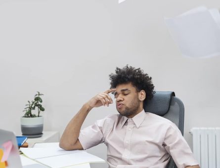 A man in a business suit sits at his desk with his head resting on his open laptop, surrounded by paperwork and office supplies, including scattered pens, notebooks, and a coffee mug. His posture is slumped, and his expression is one of sheer exhaustion and burnout, with dark circles under his eyes. The cluttered desk and dim office lighting further emphasize the stressful work environment, indicating a pressing need for better brain health and improved work-life balance. The scene poignantly captures the toll of overwork and the importance of adopting healthier habits for mental and physical well-being.