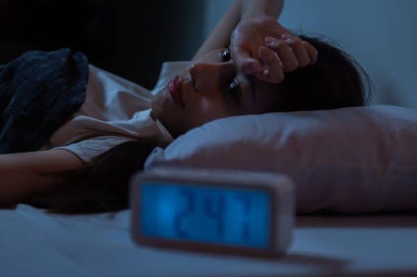 Woman laying in bed on her back, with her hand resting on her head in her hands. The clock beside her reads 2:47 am. She's wondering, "How much CBD do I need to take for sleep?"