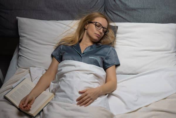 A woman wearing glasses in bed falls asleep on her bed after reading the book that's pictured laying open beside her.